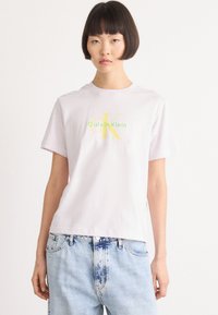 Hvit bomulls T-skjorte med en grønn og gul Calvin Klein-logo på brystet. Kombinert med lyseblå denimjeans.