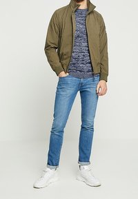 Giacca bomber verde oliva, maglione in maglia blu navy, jeans azzurri e sneakers bianche; un outfit casual con una vestibilità moderna.