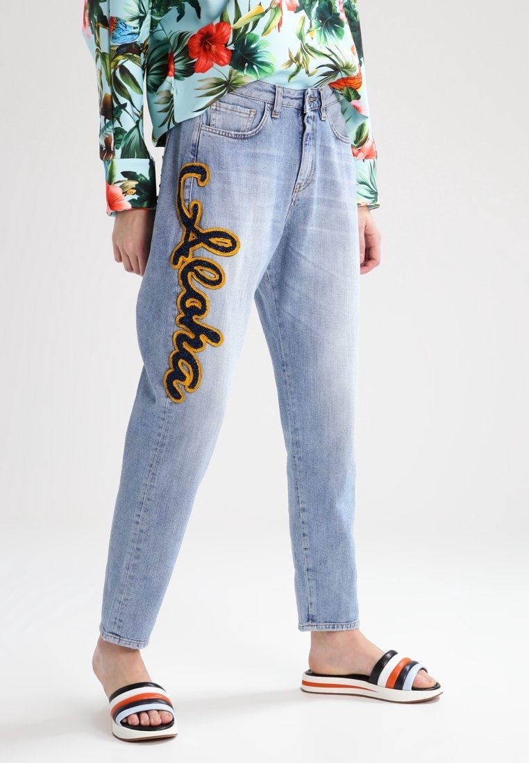 Jeans de mezclilla azul claro con "Aloha" bordado en amarillo en el lateral. Llevados con una blusa de estampado floral y sandalias de rayas múltiples.