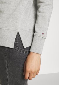 Grå sweatshirt med ribbade muddar och sidoslitsar. Har en liten logotyp på mudden. Bärs över mörkgrå jeans.