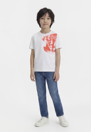 Ragazzo in piedi, indossa una maglietta bianca con stampa rossa, jeans blu e scarpe da ginnastica bianche, su sfondo bianco semplice.