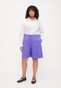 Pantaloni corte viola con una vestibilità rilassata, caratterizzati da tasche laterali, chiusura con bottone e orlo arrotolato. Abbinati a una camicia bianca a maniche lunghe.