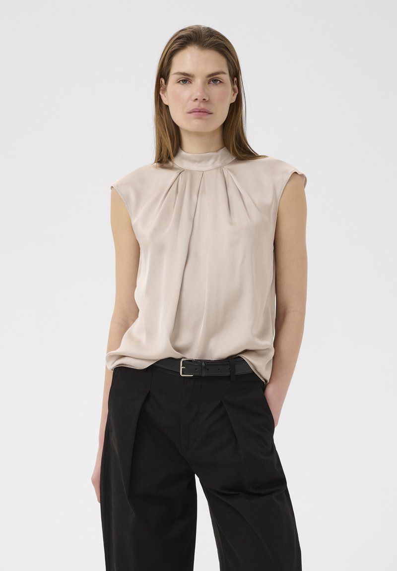Femme aux cheveux châtain clair et lisses portant un blouse beige sans manches rentrée dans un pantalon noir taille haute avec une ceinture noire.