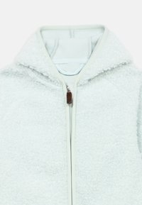 Veste polaire bleu clair avec une finition douce et texturée, dotée d'une fermeture éclair et d'une capuche. Accentuée d'une languette marron sur la fermeture éclair.