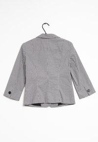 Blazer a quadri bianco e nero con maniche a tre quarti, polsini con un solo bottone e taglio su misura, appeso a un gancio nero su sfondo bianco.