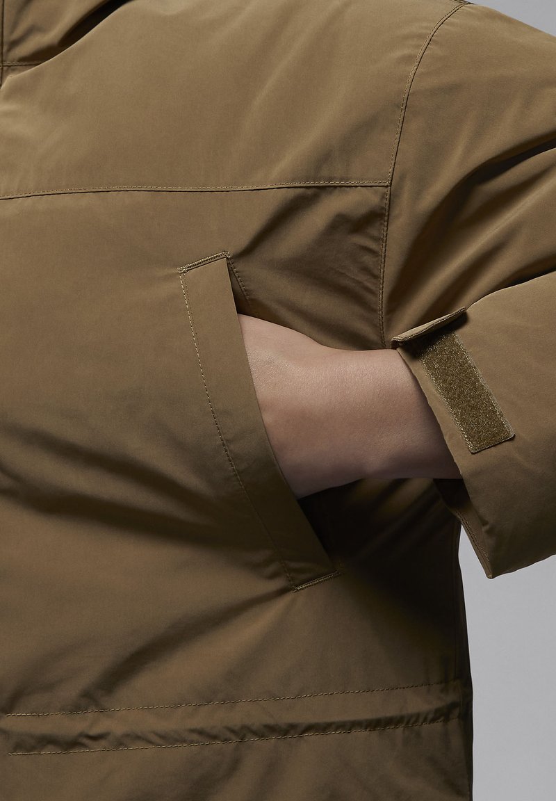 Chaqueta marrón, texturada, con un bolsillo con cremallera en la manga izquierda y una correa de Velcro en el puño. Presenta un ajuste relajado y un diseño sin costuras.