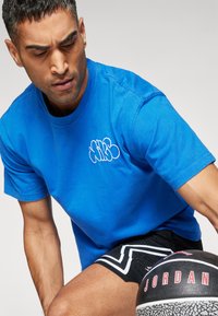 Camiseta de algodón azul con mangas cortas y cuello redondo que presenta un logo bordado en blanco. Pantalones cortos deportivos negros con acentos en zigzag en blanco.