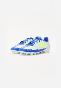 Crampons de soccer bleu et blanc avec une finition texturée, présentant des accents jaunes et un design basse. Chevilles de traction sur la semelle extérieure.