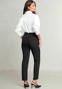 Donna in una camicia bianca con colletto e pantaloni neri su misura si volta mostrando un outfit elegante e tacchi neri affusolati su uno sfondo neutro.
