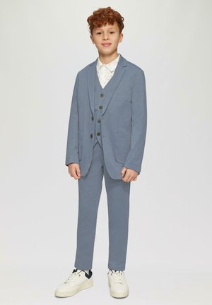 Costume bleu clair avec un gilet, une veste et un pantalon assorti. Chemise blanche avec de petits motifs noirs. Baskets blanches. Tissu lisse, coupe ajustée.