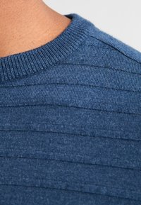 Prim-plan al unui pulover tricotat albastru, cu guler rotund și fante, prezentând un design texturat cu linii orizontale și un aspect moale.