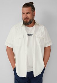 Witte shirt met korte mouwen en twee voorzakken, van textuurstof, gedragen over een ronde hals witte t-shirt met blauw logo, gecombineerd met donkere broek.