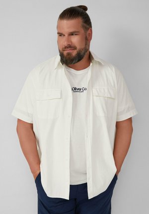 Witte shirt met korte mouwen en twee voorzakken, van textuurstof, gedragen over een ronde hals witte t-shirt met blauw logo, gecombineerd met donkere broek.