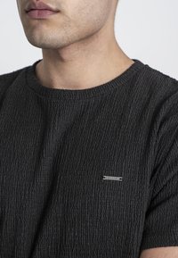 Svart texturerad t-shirt med ribbat mönster, rund halsringning och en liten metall-logotags på bröstet.