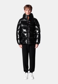 Doudoune noire brillante avec un col haut, une fermeture éclair à l'avant et des accents orange. Portée avec un pantalon noir et des baskets blanches.