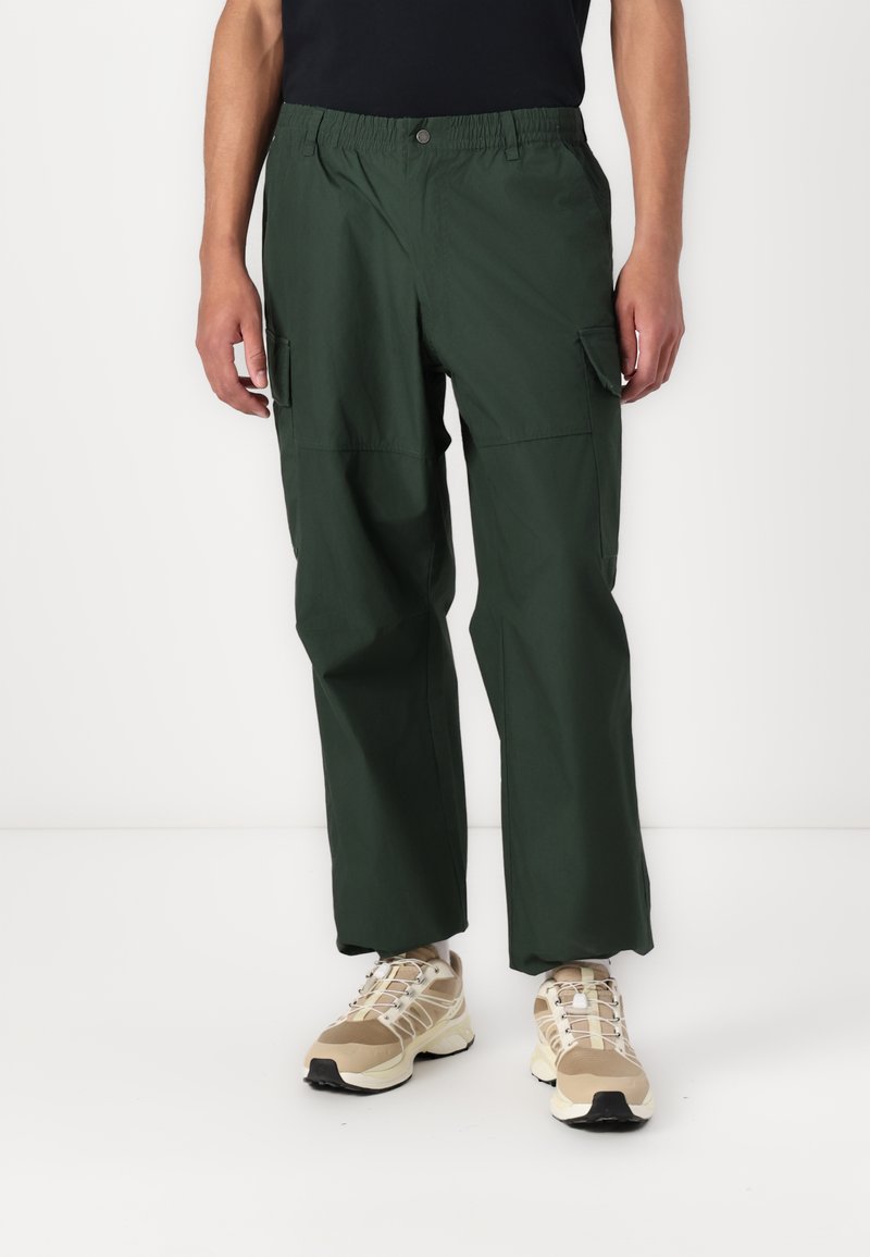 Mørkegrønne cargo-bukser har elastisk midjebånd, sidelommer og en avslappet passform. Brukt med lyse sneakers.