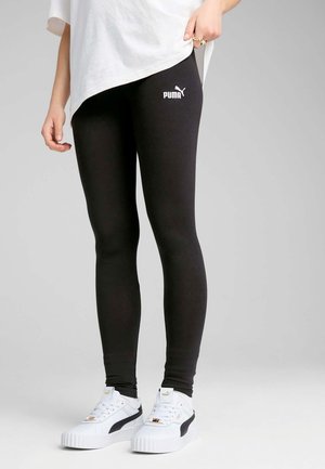 Beine in schwarzen Puma Leggings und weißen Puma Sneakers, kombiniert mit einem weißen oversized T-Shirt vor einem einfarbigen grauen Hintergrund.