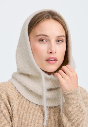 Femme portant un pull en tricot beige et une capuche douce assortie, tenant le cordon de la capuche près de son visage sur un fond clair uni.