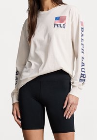 Långärmad beige bomullsskjorta med amerikansk flagga och "POLO"-logotyp på bröstet; ihop med svarta sportiga cykelbyxor.