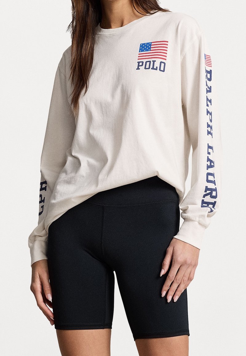 Långärmad beige bomullsskjorta med amerikansk flagga och "POLO"-logotyp på bröstet; ihop med svarta sportiga cykelbyxor.