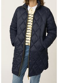 Persona che indossa una giacca imbottita blu scuro sopra un maglione a righe, in piedi con le mani in tasca, su uno sfondo chiaro.