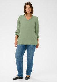 Grünes, strukturiertes Oberteil mit V-Ausschnitt und Rüschendetails, kombiniert mit blauen Jeans und schwarzen Ballerinas. Lockere Passform mit dreiviertel Ärmeln.