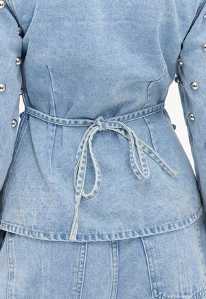 Veste en jean bleu clair avec ceinture nouée à la taille et clous métalliques argentés sur les manches, vue de dos et au niveau de la taille.