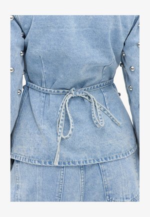 Veste en jean bleu clair avec ceinture nouée à la taille et clous métalliques argentés sur les manches, vue de dos et au niveau de la taille.