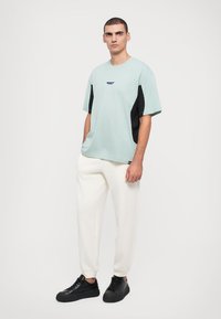 Μανίκι κοντό μπλουζίτσα σε μέντα χρώμα με μαύρες πλαϊνές ρίγες και κεντημένο λογότυπο, συνδυασμένη με λευκές φόρμες και μαύρα αθλητικά παπούτσια.