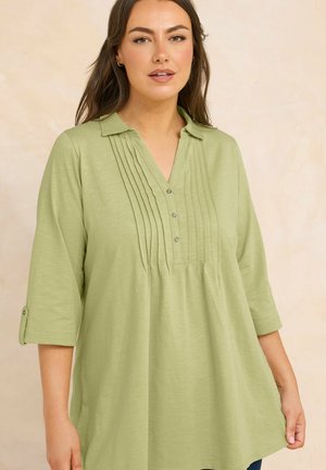 Frau trägt eine hellgrüne Tunika mit Kellerfalte-Details und 3/4-Ärmeln, hält den Saum vor beigem Hintergrund.