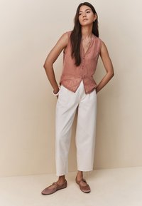 Top senza maniche color pesca con bottoni e tessuto strutturato, abbinato a pantaloni larghi color crema e ballerine marroni, in piedi di fronte a uno sfondo neutro.