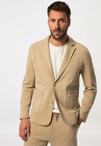 Blazer beige realizzato in morbido tessuto con due tasche frontali, rever a scollo a V e chiusura con bottone. Abbinate a pantaloni beige coordinati e una camicia bianca.