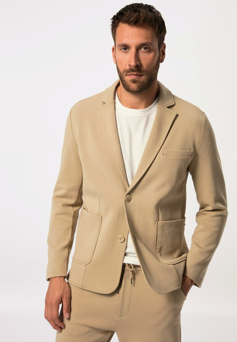 Blazer beige realizzato in morbido tessuto con due tasche frontali, rever a scollo a V e chiusura con bottone. Abbinate a pantaloni beige coordinati e una camicia bianca.