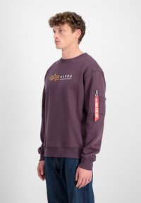 Ljubičasti dukserica od mješavine pamuka, s logom na grudima i džepom na rukavu sa zatvaračem i crvenom oznakom.