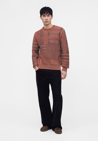 Un homme se tient debout portant un pull tricoté à motifs orange et marron, un pantalon noir à jambes larges et des chaussures marron sur un fond blanc uni.