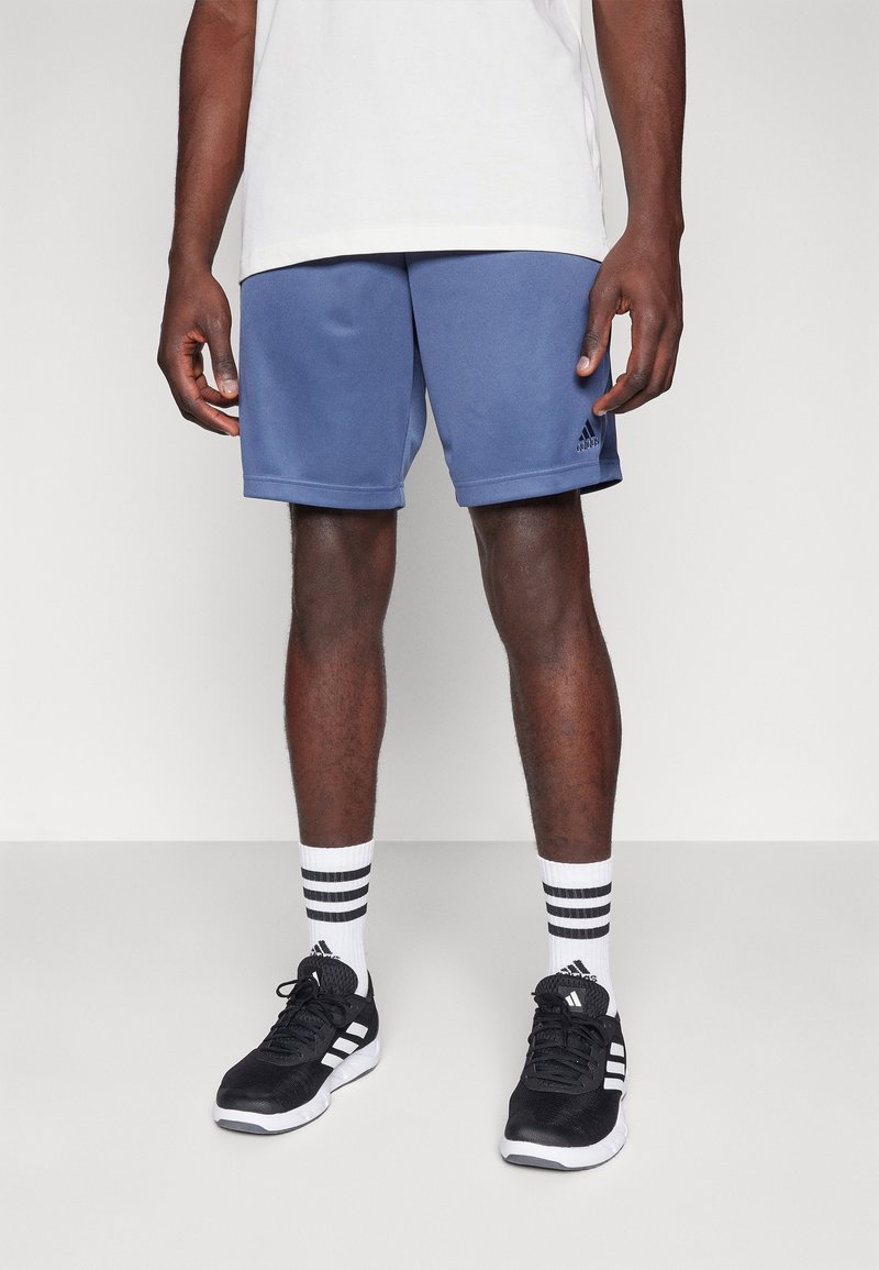 Muž v modrých šortkách Adidas, bílých ponožkách s černými pruhy a černých teniskách Adidas, stojící na světlém jednobarevném pozadí.