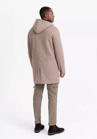 Abrigo de mezcla de lana beige con capucha, con un corte recto. Lleva encima pantalones cargo en tono beige claro y botas de tobillo marrón oscuro.