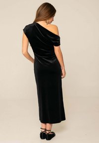 Vestido de terciopelo negro con diseño asimétrico con hombros descubiertos, silueta ajustada y dobladillo a la altura del tobillo. Textura suave y brillo sutil.