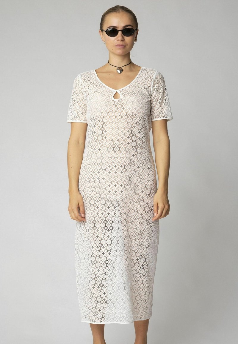Rochie midi albă din dantelă, cu mâneci scurte, model cu diamante, detaliu în formă de cruce la decolteu și croială lejeră, purtată cu ochelari de soare negri.