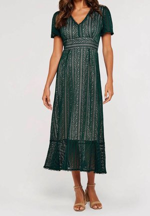 Donna che indossa un abito midi in pizzo verde scuro con maniche corte e scollo a V, abbinato a sandali beige con tacco alto.