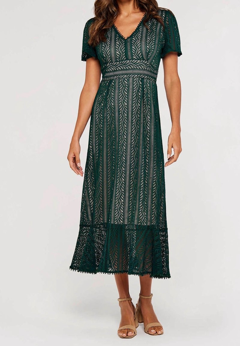 Femme portant une robe midi en dentelle vert foncé avec manches courtes et col en V, associée à des sandales à talons hauts beiges.