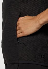 Zwarte sweatshirt met textuur, voorzien van een zijzak met een trekkoord, vertonend een subtiel zigzagpatroon en een geborduurd "R"-logo.