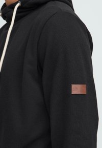 Musta huppari, joka on valmistettu puuvillasekoitteesta, jossa on beige naru ja ruskea nahkainen logopatch vasemmassa hihassa.