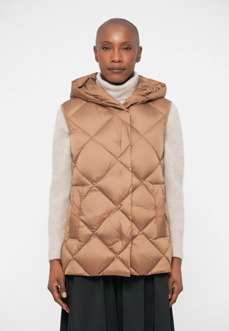 Gilet matelassé brun avec capuche, présentant une texture lisse et un grand motif en losanges brodé. Associé à un pull léger et une jupe sombre.