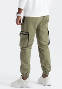Pantalons cargo en tissu vert olive avec des poignets élastiques. Caractérisés par deux poches zippées noires sur les côtés et un minimum de détails de couture.