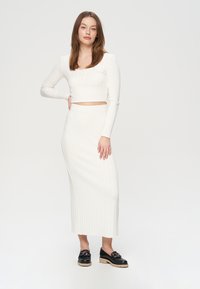 Balts ribots adīts cropped tops ar garām piedurknēm, savienots ar atbilstošu midi garuma ribotu svārku. Modelis valkā melnas kurpes ar masīvu zoli.