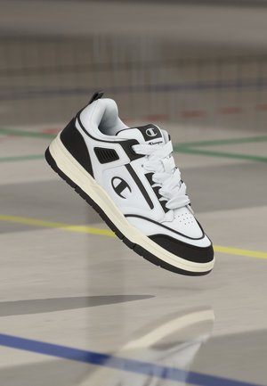 Baskets Champion blanches et noires avec lacets flottant au-dessus d’un sol de gym marqué de lignes sportives de plusieurs couleurs.