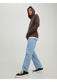 Sweat à capuche marron avec poche avant, porté sur une chemise blanche, associé à un jean bleu clair et des baskets noires.