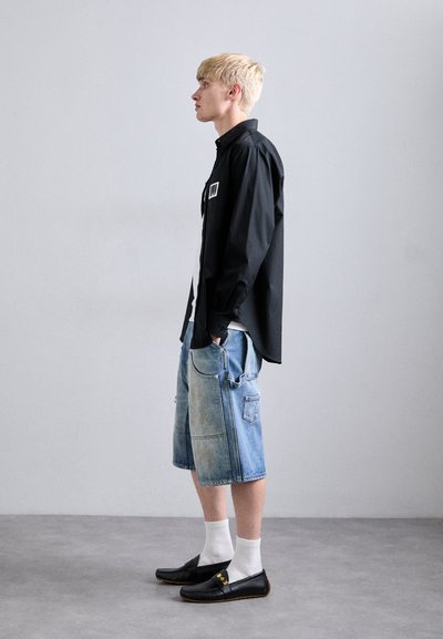 Ung man iklädd svart långärmad tröja, långa denimshorts, vita strumpor och svarta slip-on skor, står på sidan mot en ren vägg.