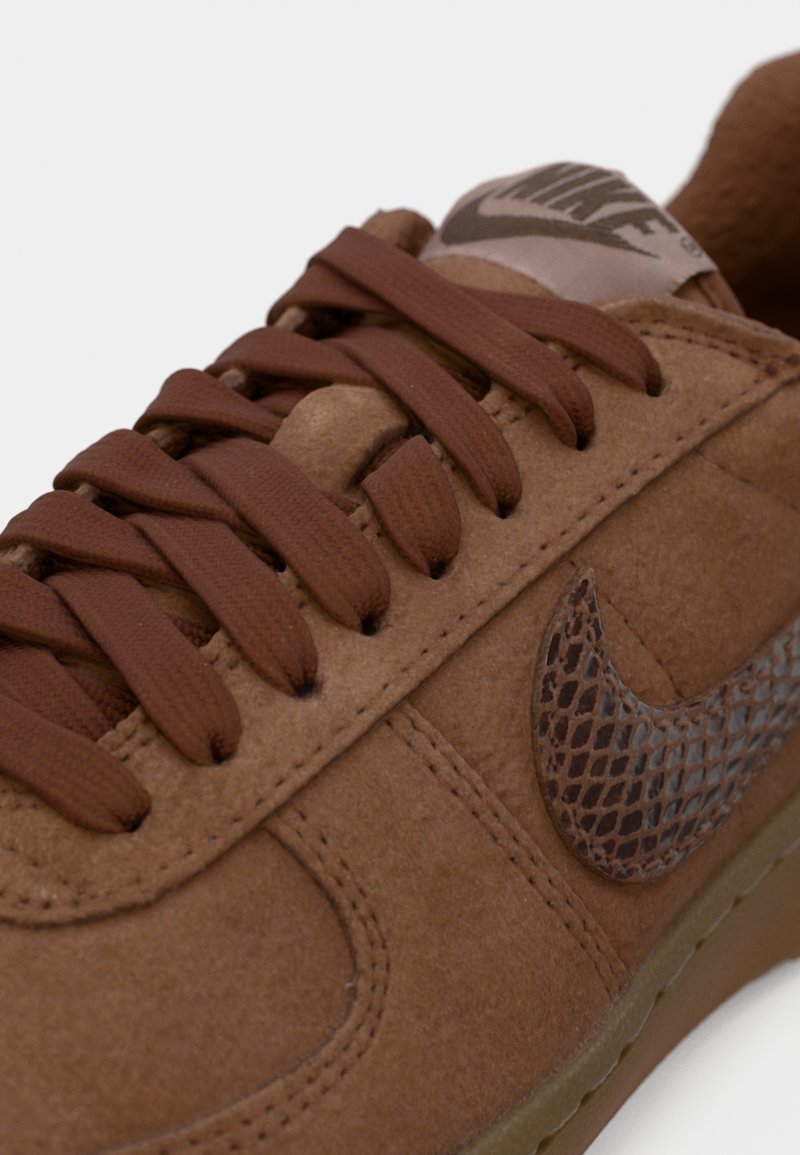 Braune Wildledersneaker mit strukturiertem Seiten-Swoosh, runden braunen Schnürsenkeln und einer gepolsterten Zunge mit einem Logo-Patch. Flache Gummisohle.